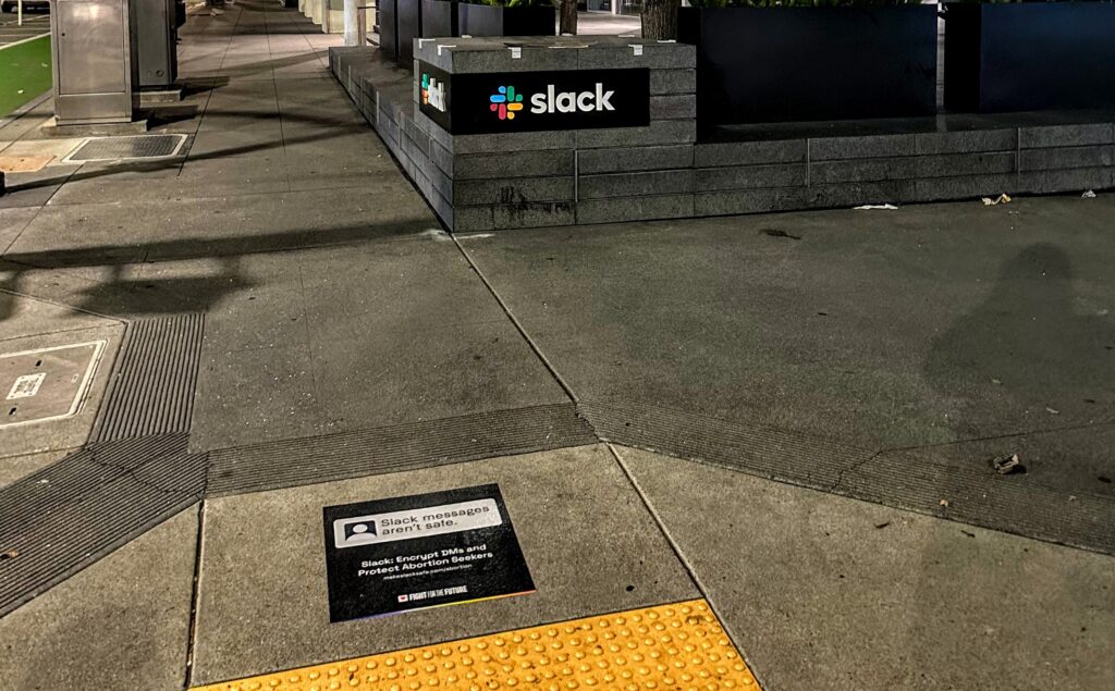 Sidewalk decal with text "Slack Messages Aren't Safe. Slack: Encrypt DMs and Protect Abortion Seekers makeslacksafe.com/abortion"; Slack logo in background