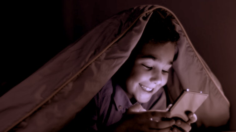 smiling kid playing game on phone under blanket at night